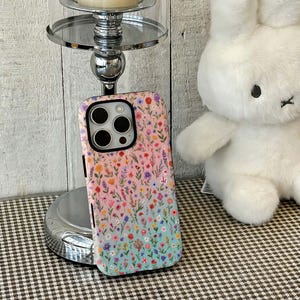 May include: A floral phone case with a gradient design. The case features a colorful array of flowers in shades of pink, purple, blue, and yellow against a light blue and pink background. The case is designed for a smartphone with multiple camera lenses.