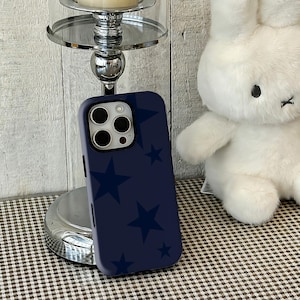 以下が含まれることがあります： 濃紺の携帯電話ケース。濃い青色の星模様が特徴です。白いぬいぐるみウサギと銀色のキャンドルホルダーの隣に表示されています。携帯電話ケースにはカメラ用の切り欠きがあります。