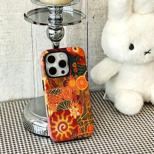 May include: A rectangular smartphone case featuring a vibrant collage of warm-toned graphics, including orange slices, stylised suns, floral patterns, and butterflies in shades of red, orange, yellow, and deep green, with a black camera cutout.