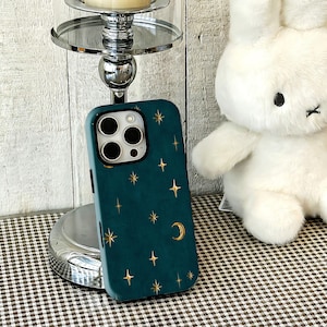 May include: A dark teal smartphone case featuring a celestial pattern of gold-coloured stars and a crescent moon. The case has a cutout for a triple-lens camera system and covers the back and sides of the device with a smooth, matte-like finish.