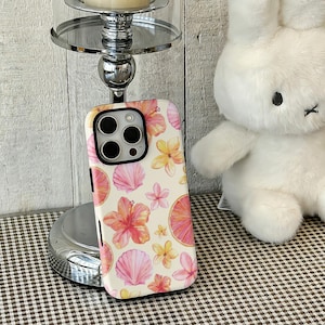 May include: A phone case with a tropical floral pattern. It features pink hibiscus flowers, yellow flowers, and pink seashells on a cream background. The case has a black border and a camera cutout.