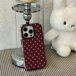 Puede incluir: Funda de teléfono burdeos con un patrón de lunares rosas. La funda tiene un recorte para la cámara. También se ve un conejito de peluche blanco y un candelabro plateado.