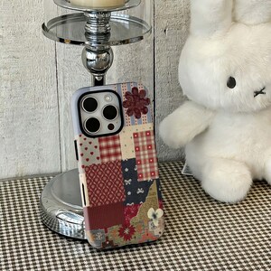 Könnte beinhalten: Eine Smartphone-Hülle mit Patchwork-Design, bestehend aus verschiedenen Mustern wie rot-weißem Karomuster, kleinen Herzen, Blumenmotiven und geometrischen Formen in Rot-, Blau- und Cremetönen, mit einer Aussparung für ein Dreifach-Kamerasystem.