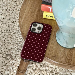 Puede incluir: Una funda de tel&eacute;fono burdeos con un patr&oacute;n de peque&ntilde;os corazones rosas. La funda est&aacute; sobre una superficie de madera, junto a una pila de libros y un objeto decorativo. La funda del tel&eacute;fono tiene un recorte para la c&aacute;mara.