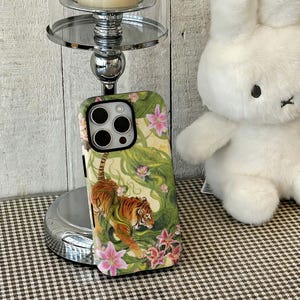 May include: A rectangular smartphone case featuring a detailed illustration of an orange and black tiger walking through lush green foliage and pink lily flowers. The case has a black camera cutout frame and a smooth, glossy finish on the back surface.