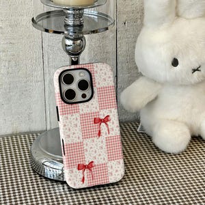 May include: A pink and white patchwork phone case with a floral and gingham pattern, accented with red bows. The case is designed for a smartphone and features a camera cutout.