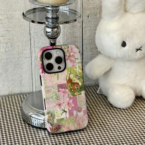 May include: A floral phone case with a patchwork design featuring pink lilies and a tiger illustration. The case is shown next to a silver candle holder and a white stuffed bunny.