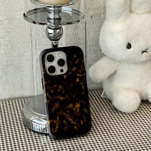 Puede incluir: Funda de teléfono negra y marrón con estampado de carey. La funda tiene un acabado brillante y un recorte para la cámara. Un conejito de peluche blanco está en el fondo. Un candelabro plateado también está en el fondo.