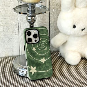 May include: A forest green smartphone case featuring a hand-drawn white spiral design with scattered stars. The case has a black camera cutout frame and a textured, matte finish, designed to fit a device with a triple-lens camera system.