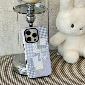 May include: A light blue and white patchwork phone case with a variety of patterns, including gingham, floral, and striped designs. The case is shown on a silver stand with a candle and next to a white stuffed bunny.