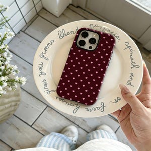 Puede incluir: Funda de tel&eacute;fono burdeos con un patr&oacute;n de peque&ntilde;os corazones rosas. La funda est&aacute; sobre un plato blanco con texto manuscrito. Accesorio protector para smartphone.