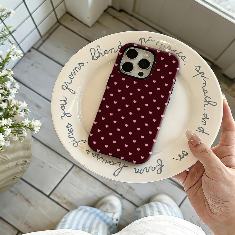 Puede incluir: Funda de tel&eacute;fono burdeos con un patr&oacute;n de peque&ntilde;os corazones rosas. La funda est&aacute; sobre un plato blanco con texto manuscrito. Accesorio protector para smartphone.