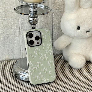 May include: A light green phone case with a white floral pattern. The case is shown next to a white stuffed bunny and a silver candle holder. The phone case has a camera cutout and black accents.