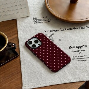 Puede incluir: Una funda de tel&eacute;fono burdeos con un patr&oacute;n de peque&ntilde;os corazones rosas. La funda est&aacute; sobre una superficie de lino blanco con texto en franc&eacute;s y un calendario. Tambi&eacute;n se ve una taza de caf&eacute; marr&oacute;n oscuro.