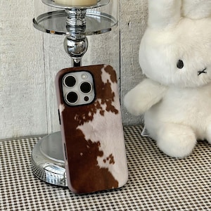 May include: A brown and white cowhide-patterned phone case. The case has a glossy finish and a camera cutout. A white stuffed bunny is in the background. The phone case is next to a silver candle holder.