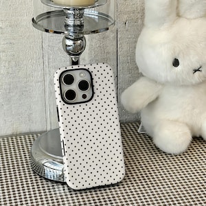 Può includere: Custodia per telefono bianca con pois neri. La custodia ha un bordo nero e un ritaglio per la fotocamera. Un coniglietto bianco di peluche è sullo sfondo, insieme a un candelabro argentato. La custodia è su una superficie a scacchi marrone e bianca.