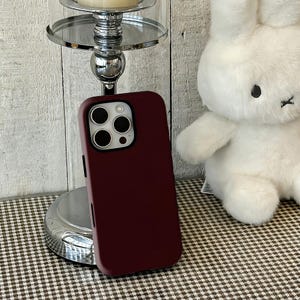 Może przedstawiać: Bordowa obudowa telefonu z wycięciem na aparat, spoczywająca na srebrnym stojaku. Biały pluszowy królik jest w tle. Obudowa telefonu jest w jednolitym kolorze i wydaje się być wykonana z gładkiego materiału.