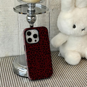May include: A red and black leopard print phone case. The case has a raised edge around the camera and a smooth, glossy finish. The case is shown next to a white stuffed bunny and a silver candle holder.