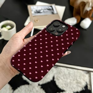 Puede incluir: Una funda de tel&eacute;fono burdeos con un patr&oacute;n de peque&ntilde;os corazones rosas. La funda tiene un recorte negro para la lente de la c&aacute;mara y est&aacute; sujeta con una mano. El fondo incluye una taza blanca, un libro y un peluche.