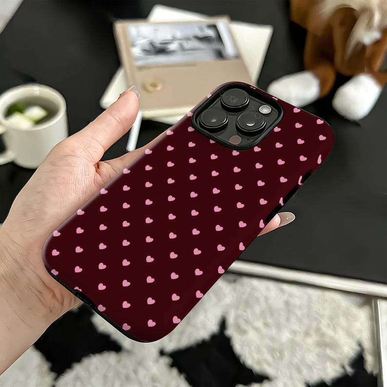 Puede incluir: Una funda de tel&eacute;fono burdeos con un patr&oacute;n de peque&ntilde;os corazones rosas. La funda tiene un recorte negro para la lente de la c&aacute;mara y est&aacute; sujeta con una mano. El fondo incluye una taza blanca, un libro y un peluche.