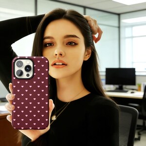 Puede incluir: Una funda de tel&eacute;fono burdeos con un patr&oacute;n de peque&ntilde;os corazones rosas. La funda tiene un borde negro y recortes para la c&aacute;mara y los botones. La funda del tel&eacute;fono se sostiene frente a una persona.