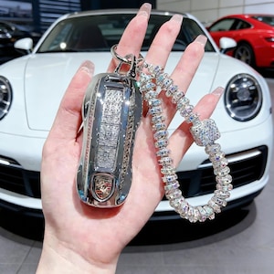 Puede incluir: Un llavero plateado de coche Porsche, adornado con cristales brillantes. El llavero está unido a una correa de muñeca con cristales, creando un accesorio glamuroso. El fondo presenta un Porsche blanco.