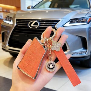 Puede incluir: Funda de llave de coche de cuero naranja con detalles dorados y una correa a juego. El llavero tiene símbolos de llave y botones de entrada sin llave. Un llavero dorado con un trébol de cristal y un dije circular está adjunto.