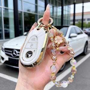 May include: White and gold car key fob with a decorative beaded keychain. The key fob has a gold trim and a diamond-like border. The keychain features iridescent beads, gold accents, and butterfly charms.