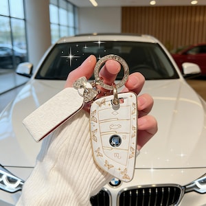 May include: White and gold car key fob cover with butterfly and floral designs. The fob has a gold-colored metal ring with a diamond-like encrusted circle. The key fob is paired with a white leather key tag.