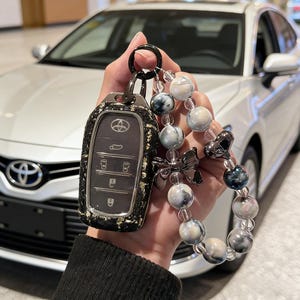 May include: A black and gold patterned car key fob cover with a beaded keychain. The keychain features clear beads, marbled grey and white beads, and a silver bow charm. The car in the background is silver.