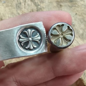 Puede incluir: Un brazalete de metal plateado con un emblema circular que presenta un diseño de cruz de cuatro pétalos en relieve y una herramienta de sello circular a juego con un patrón de cruz hundido para crear el detalle en relieve del brazalete.
