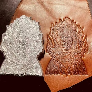 May include: A clear acrylic stamp and a leather impression of a detailed, ornate face with a fiery headdress. The leather impression is a rich brown color, showcasing the intricate design. The stamp is next to the leather.