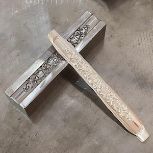 Könnte beinhalten: Ein silbernes Metallwerkzeug mit kunstvollen Blumen- und Rankenmustern. Das Werkzeug liegt diagonal auf einer rechteckigen Metallform mit ähnlichen Gravuren. Der Hintergrund ist eine strukturierte, metallische Oberfläche.