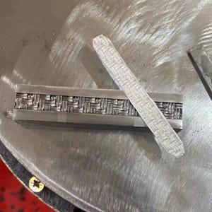 Könnte beinhalten: Zwei silberne Metallstäbe mit einem gewebten Muster. Einer ist rechteckig, der andere ein langes, dünnes Rechteck. Die Stäbe liegen auf einer silbernen Metalloberfläche.