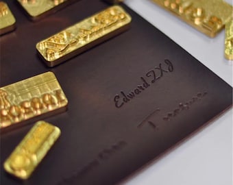 May include: A collection of gold-colored metal stamps arranged on a dark brown leather surface. The stamps have detailed relief designs. The leather is embossed with the words "Edward ZxJ Treasure".