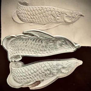 May include: Three embossed fish designs, showing detailed scale patterns and fins. The fish are in different stages of completion. The designs are in a neutral colour palette, with varying levels of relief, possibly made from leather or clay.