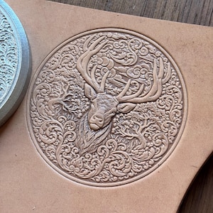 May include: A circular leather stamp depicting a stag's head with elaborate antlers, set within a detailed forest scene. The stamp is pressed into light brown leather, displaying the embossed design. The design is approximately 10 cm in diameter.