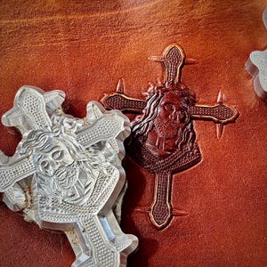 Puede incluir: Primer plano de una superficie de cuero marrón con dos cruces. Una cruz está grabada en un marrón más oscuro, representando una figura religiosa. La otra es un molde de plástico transparente del mismo diseño. El fondo es de un marrón rojizo texturizado.
