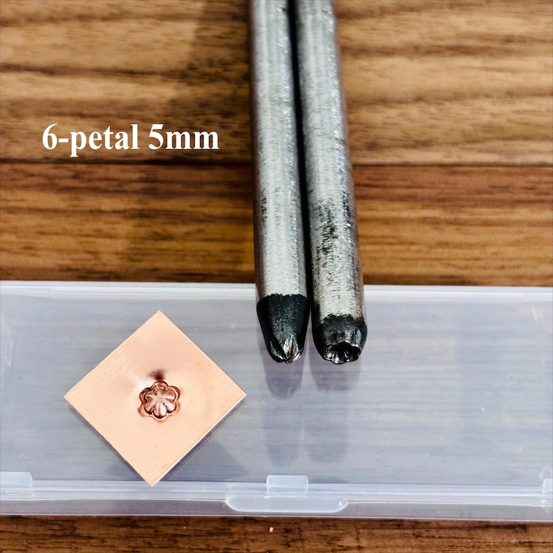 May include: Metal stamping tools with a 6-petal 5mm design. Two silver metal tools are positioned next to a small copper square with a stamped flower design. The text "6-petal 5mm" is visible in the image.