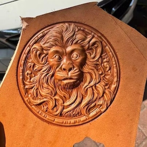 May include: A detailed, hand-tooled leather piece featuring a lion's head in a circular design. The lion's mane and facial features are intricately carved, showcasing the natural tan color of the leather. The edges of the leather are visible, adding to the handcrafted aesthetic.