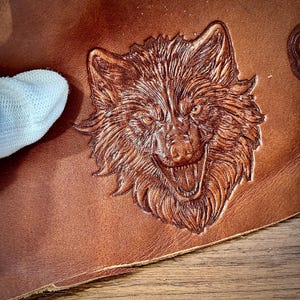 May include: Close-up of a brown leather item with a detailed embossed wolf face. The wolf's features are intricately carved, showcasing its open mouth and sharp teeth. The leather has a rich, warm tone, and the carving adds depth and texture.