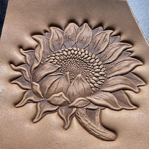 May include: Close-up of a tan leather piece with a detailed, embossed sunflower design. The sunflower is in full bloom, with textured petals and a detailed center. The leather has a subtle grain.