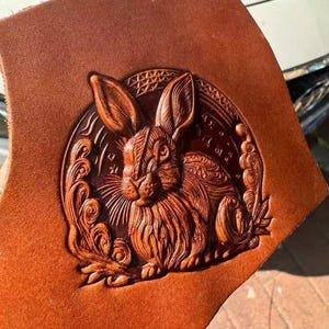 May include: Close-up of a brown leather piece with a detailed embossed image of a rabbit. The rabbit is centered within a circular design, surrounded by decorative swirls and patterns. The leather has a rich, warm tone.