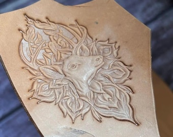 May include: A tan leather piece with a detailed embossed design of a deer's head surrounded by leaves and antlers. The deer is facing upwards, and the design is centered on the leather. The leather has a natural, unfinished look.