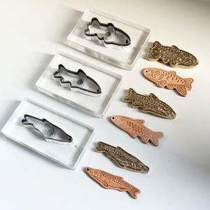 Könnte beinhalten: Eine Auswahl an fischförmigen Ausstechern und geprägten Metallfischen. Die Ausstecher sind silberfarbenes Metall mit schwarzen Umrissen, eingebettet in transparente Acrylblöcke. Die geprägten Fische sind in Gold- und Kupfertönen gehalten und zeigen detaillierte Schuppen und Flossen.