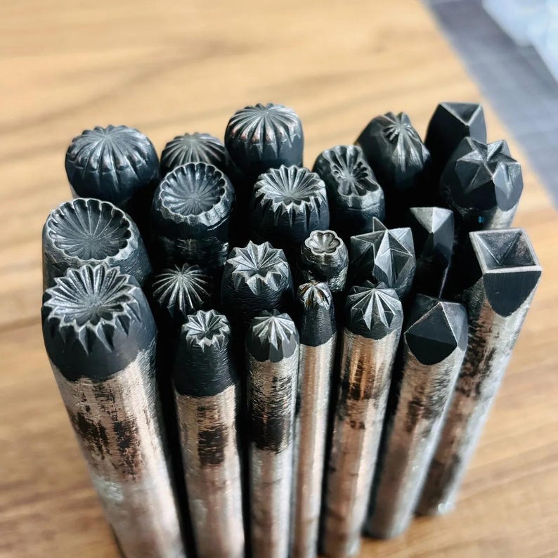 May include: A selection of metal leather stamping tools, each with a different decorative end design. The tools have a silver finish and feature detailed patterns, including floral and geometric shapes. The tools are arranged closely together.