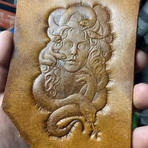 May include: A tan leather piece featuring an embossed image of a woman with snakes in her hair. The snakes are intricately detailed, and the woman's face has a serene expression. The leather has a natural, textured appearance.