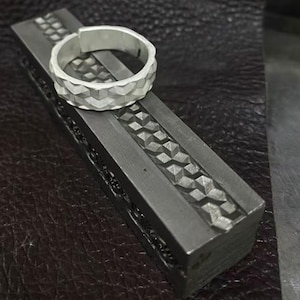May include: A silver ring with a geometric pattern rests on a dark gray metal tool. The ring has a textured surface with a series of small, angled facets. The tool has a detailed, repeating geometric design.