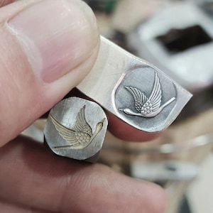 May include: Two silver metal stamps with a flying bird design. One stamp is rectangular, the other is square. The bird is depicted with outstretched wings and a long neck, with detailed feathering. The stamps are held by a person.