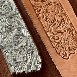 Puede incluir: Primer plano de una herramienta de metal y un trozo de cuero marrón claro, ambos con intrincados diseños florales y de volutas. La herramienta de metal es plateada, mientras que el cuero muestra detalles en relieve. Los diseños incluyen flores, hojas y patrones arremolinados.
